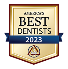 The image shows a badge with text that reads  AMERICA S BEST DENTISTS 2023  and includes a gold seal, a blue ribbon, and a graphic of a toothbrush and dental tools. It features a banner at the top with the text  BEST DENTISTS  in bold letters, followed by the year  2023  below it. The background of the badge is white, and there are two blue ribbons on either side of the seal.