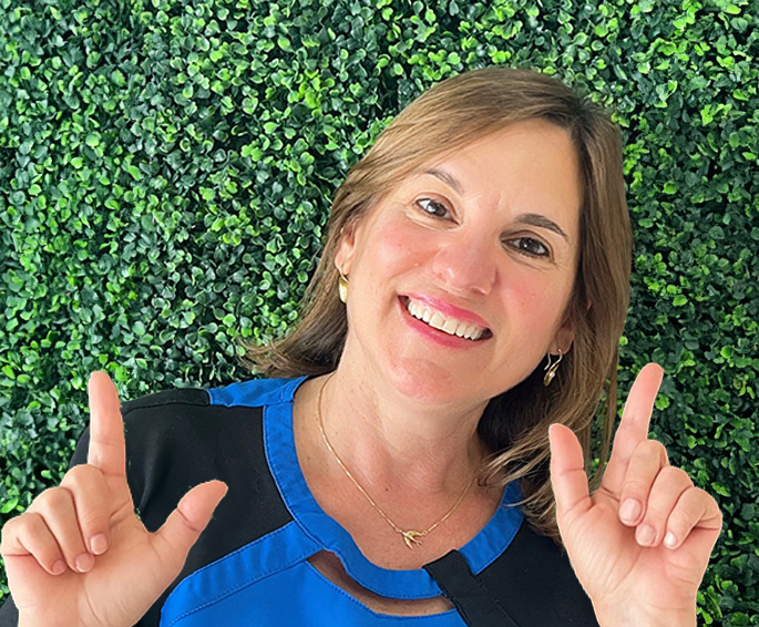The image shows a woman with a smile, making a peace sign gesture with her right hand, standing against a backdrop of green foliage.