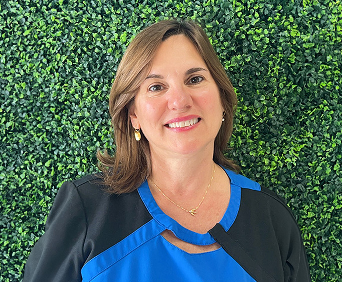 A smiling woman posing against a green wall with a white border, wearing a blue top and gold jewelry.