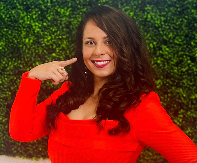 A woman with curly hair, wearing red, pointing at her nose.