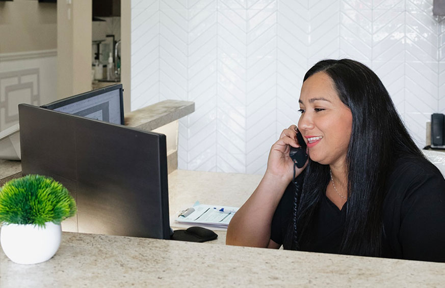 The image shows a woman seated at a desk with a computer monitor displaying a purple screen and a keyboard in front of her, working in an office environment with a window and blinds behind her.