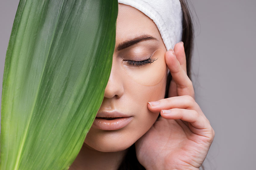 Woman with closed eyes holding a large leaf over her face, with a mask on her nose and fingers gently touching her cheek.