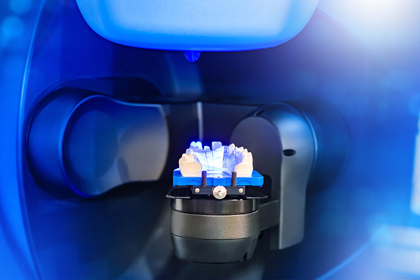 The image shows a close-up view of a blue gemstone attached to a mechanical device with a clear blue background, possibly indicating an object of value being processed or prepared for display.