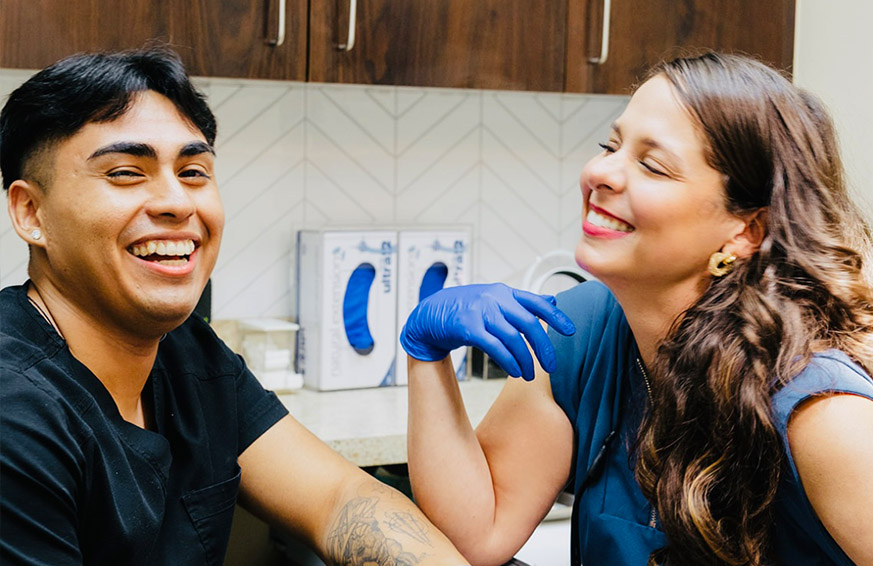 The image shows a woman sitting in a dental chair with a smile, wearing a white top, and looking directly at the camera.
