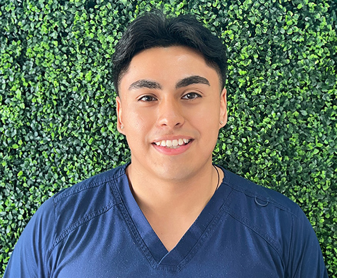 The image shows a person standing against a green wall with a smile, wearing a blue t-shirt and dark pants.