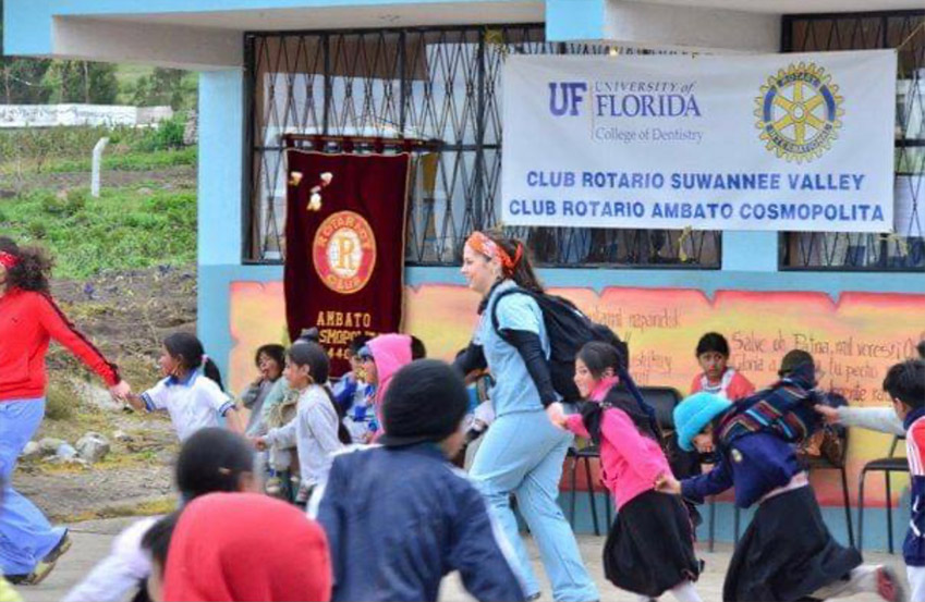 The image shows a group of people gathered outside a building with a sign that reads  UF FLORIDA CLUB ROTARIO SWAMNE VALLEY ABATEMOSCOPOLITA  in Spanish, alongside a banner for a local club and a poster advertising an event.
