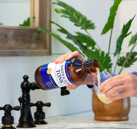 The image shows a person pouring a liquid from a bottle into a glass using a faucet, with a backdrop featuring a plant and a wooden surface.