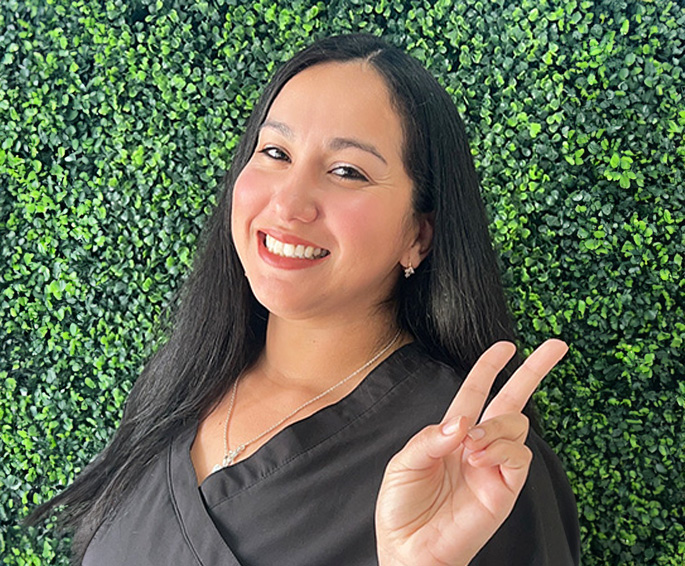 A woman with long dark hair smiles and poses peacefully with her right hand holding up a peace sign.