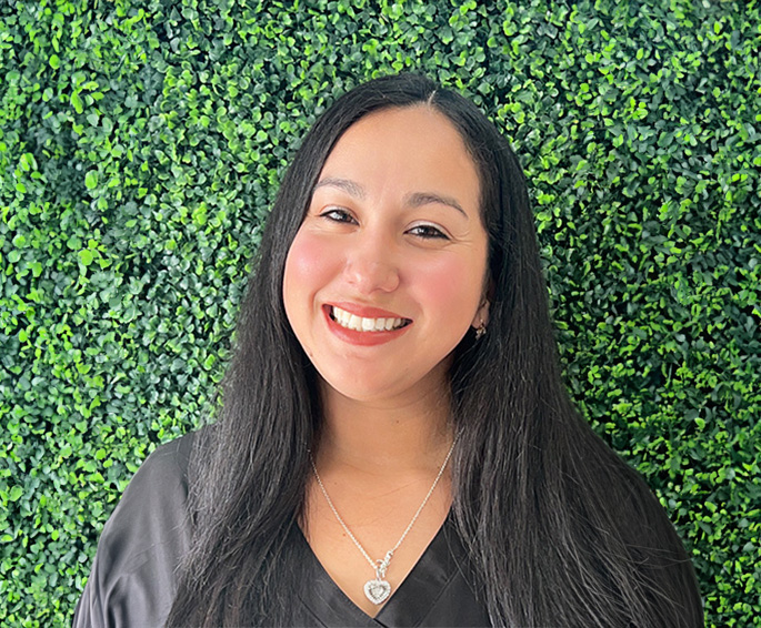 The image shows a person smiling at the camera, standing against a green backdrop with a patterned wall behind them.