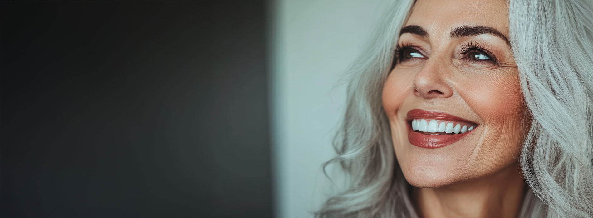 The image shows a woman with gray hair smiling at the camera, set against a blurred background with a rectangular frame on the left side.
