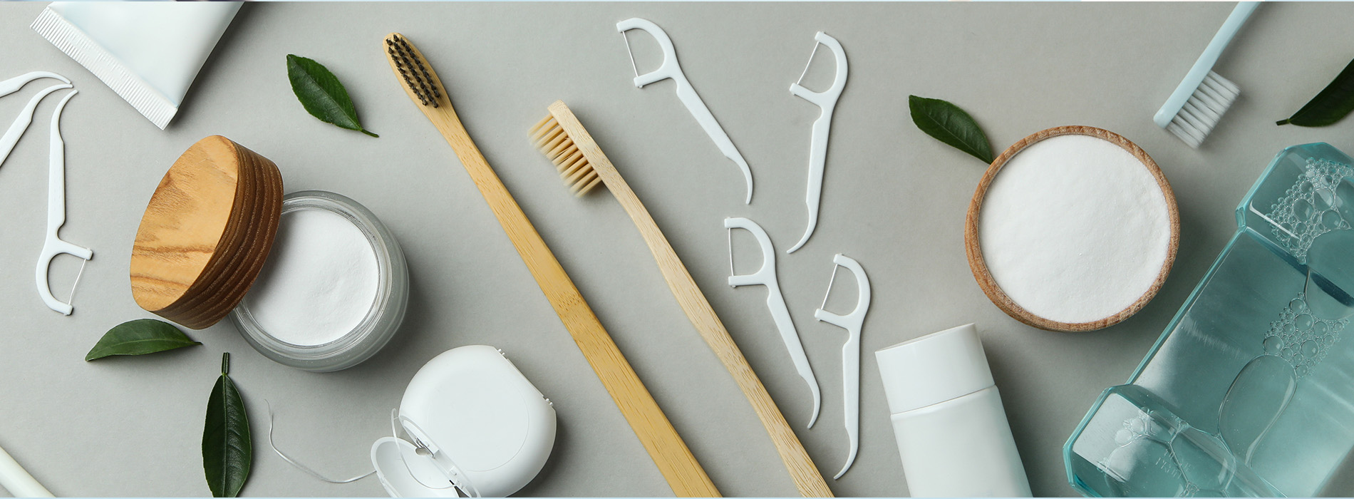 The image features a collection of beauty and skincare products arranged on a surface with a botanical theme, including wooden spoons, toothbrushes, jars, bottles, and leaves, set against a grey background.