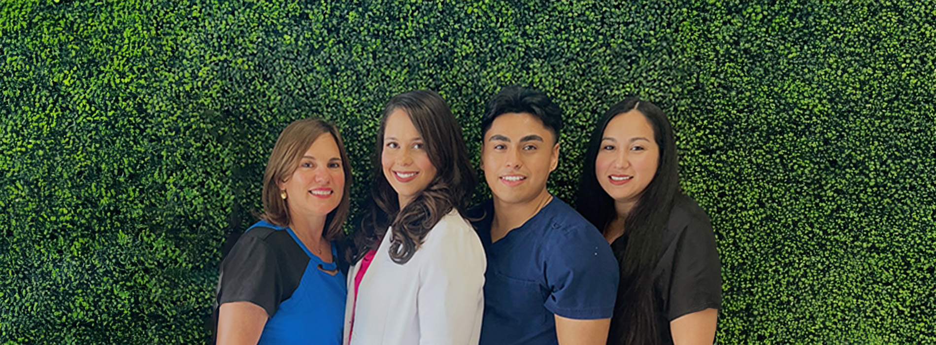 The image shows four individuals posing together in front of a backdrop with green foliage  they are smiling and appear to be at an event or gathering.