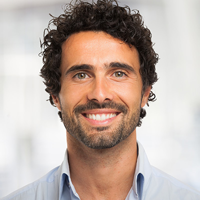 The image shows a smiling man with curly hair and facial stubble, wearing a light blue shirt, against a plain background.