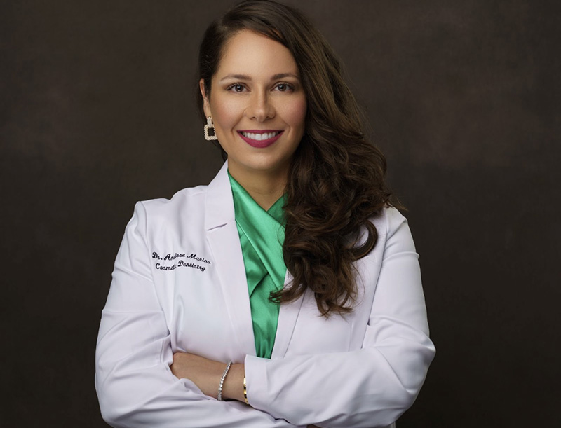 A woman in a white coat stands confidently with her arms crossed, wearing a green top and a smile.