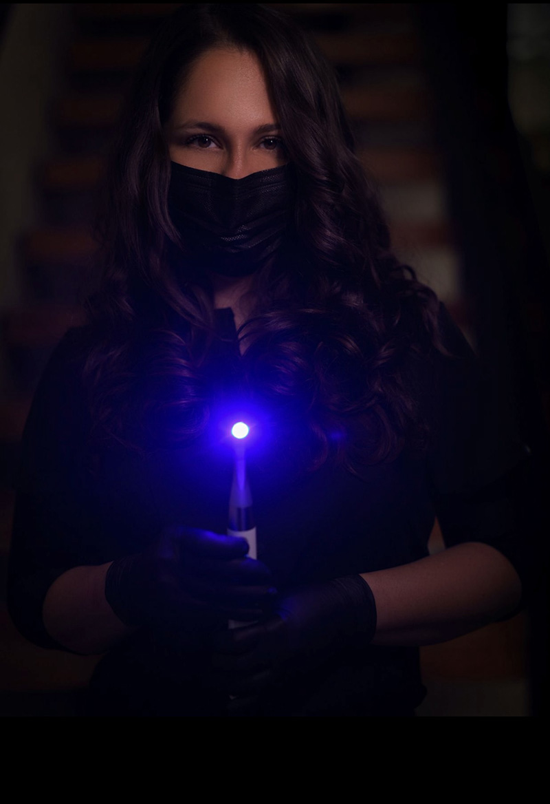 A woman wearing a black mask and gloves holds a blue object, possibly a light or device, against a dark background with wooden planks.