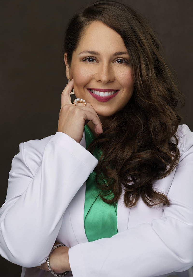 The image features a woman wearing a white coat, standing with her hands on her hips, smiling at the camera. She has long brown hair and is wearing a necklace.