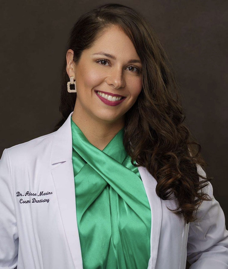 The image shows a woman wearing a white lab coat with green accents, a turquoise blouse, and a pearl necklace, standing against a dark background with a logo watermark on the upper right corner.