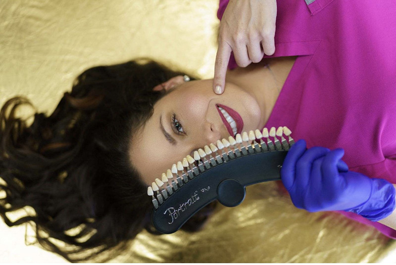 A woman with long hair lying on her back, holding a dental mirror above her face, smiling at the camera, while wearing a blue glove and a stethoscope around her neck.