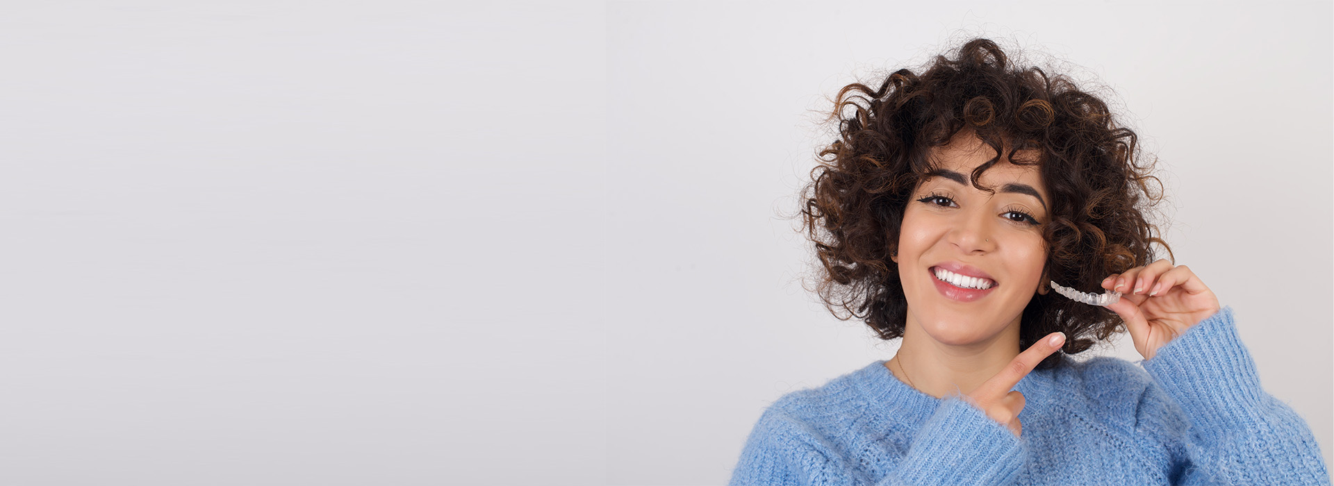 The image shows a smiling person with curly hair holding a toothbrush, set against a white background.