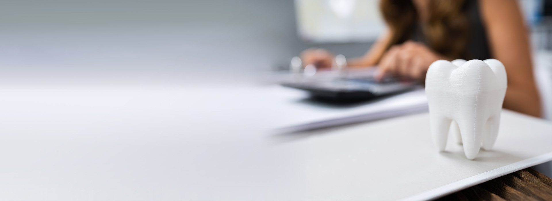 The image shows a person sitting at a desk with a laptop, papers, and a small figurine of a tooth next to them, likely indicating dental services or a dentist s office.