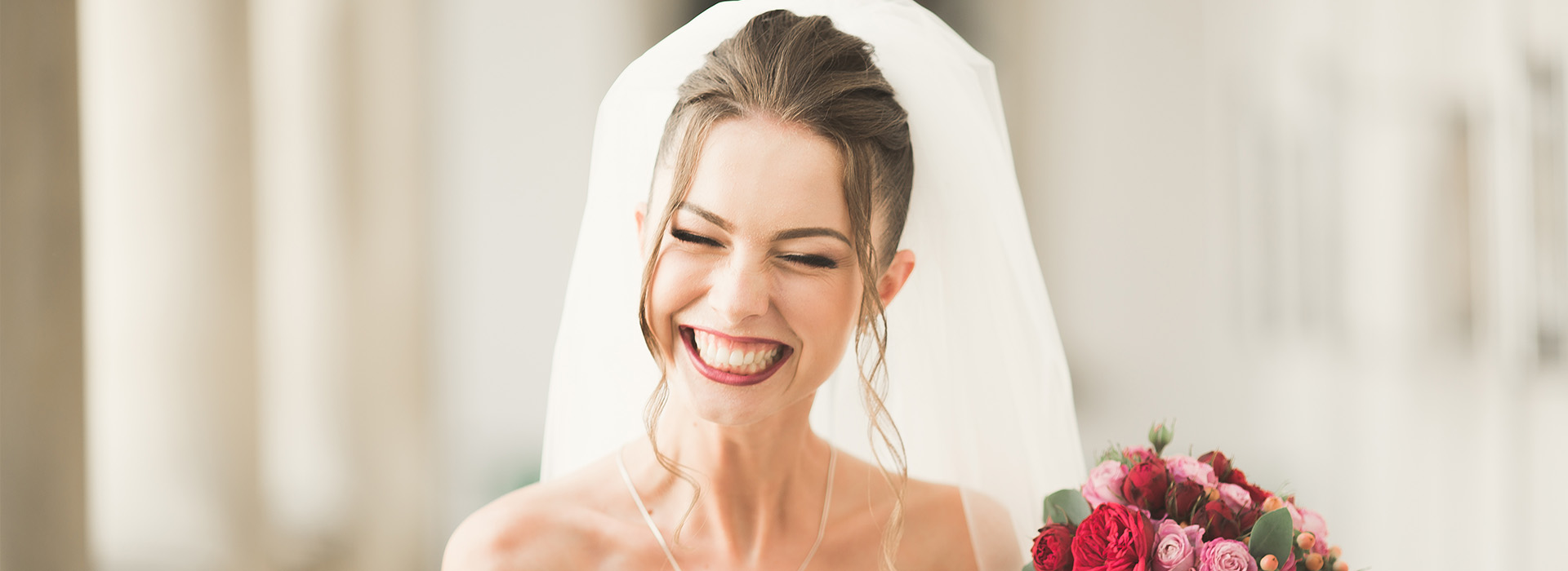 A bride wearing a wedding veil and smiling at the camera, standing next to a bouquet of flowers.