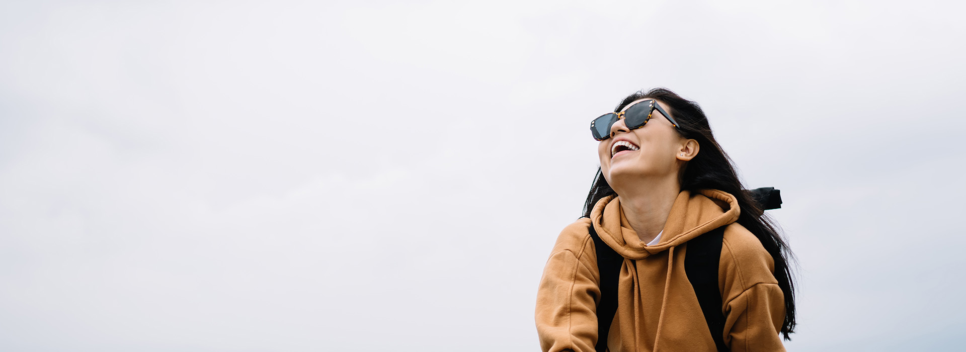 A person is standing outdoors with their back to the camera, wearing sunglasses and a backpack, against a bright sky background.