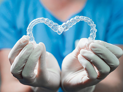 The image shows a person s hand holding a heart-shaped object with a clear plastic texture, which appears to be a dental appliance or retainer, against a background that suggests a clean environment, possibly a dental office.