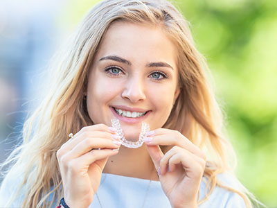 The image shows a young woman with a radiant smile, holding up a clear plastic object near her mouth, which appears to be a dental appliance, possibly an orthodontic device like a retainer or aligner, against a blurred background that suggests an outdoor setting during the daytime.