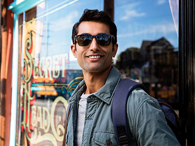 A man wearing sunglasses stands outside a storefront with a backpack, smiling at the camera.