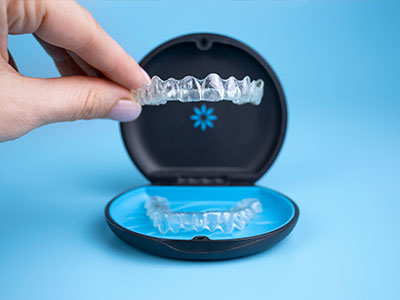 The image shows a hand holding a transparent plastic tray containing a set of clear aligners, which are part of an orthodontic treatment system, with one aligner placed inside a blue case that has the word  SMILE  on it.