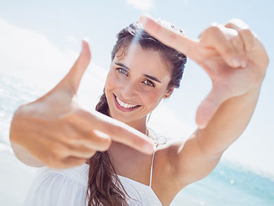 The image shows a young woman with long hair smiling at the camera while holding up two fingers to frame her face.