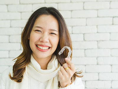 The image shows a smiling woman holding a toothbrush, with a man in the background also smiling. They both appear cheerful and are indoors with a brick wall visible behind them.